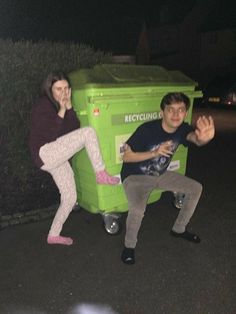 two people sitting on top of a green recycling bin