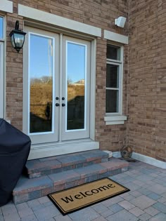 a welcome mat on the front steps of a brick building with two doors and a black umbrella