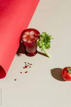 a red drink with ice and garnish next to a tomato on a white surface