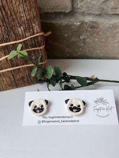 a pair of earrings with a pug face on them sitting next to a plant