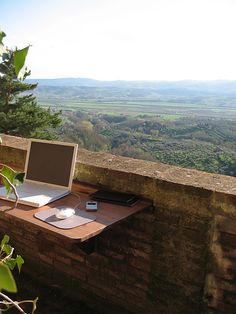 an open laptop computer sitting on top of a wooden desk next to a tree filled hillside