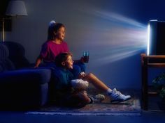 two children are sitting on the floor watching something in front of them with a flashlight