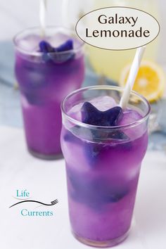 two glasses filled with purple liquid and ice on top of a white table next to lemon slices