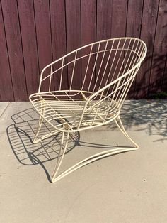 a white wire chair sitting on top of a cement ground next to a wooden fence