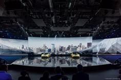 two yellow cars are on display in front of a large screen with cityscape