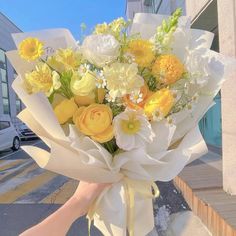 a bouquet of yellow and white flowers is being held by someone's hand on the sidewalk