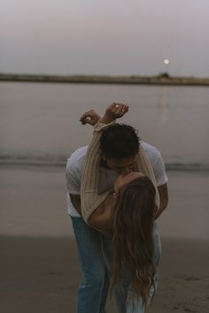 a man and woman kissing on the beach at dusk with the moon in the distance