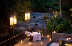 two people sitting at a table in front of a river with lit candles on it