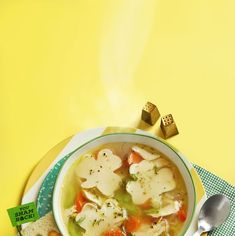 a bowl of soup with dumplings and carrots on a yellow tablecloth next to two spoons