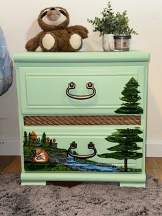 a teddy bear sitting on top of a green dresser with trees and animals painted on it