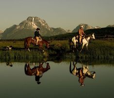 two people are riding horses in the grass near water with mountains in the back ground