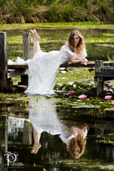 a woman in a white dress is sitting on a wooden bench by the water with her reflection