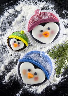 three frosted cupcakes decorated like penguins in hats and scarfs on top of snow