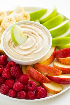 a bowl of dip surrounded by sliced bananas, raspberries and apples on a plate