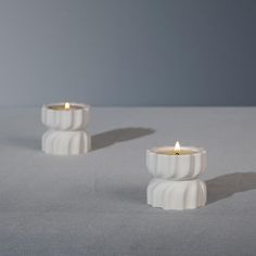 two white candles sitting next to each other on top of a gray surface with one candle lit in the middle