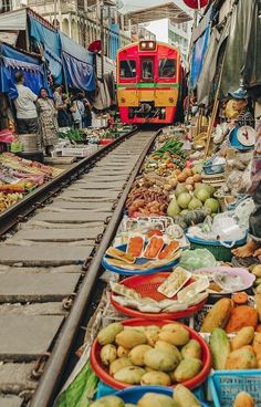 an outdoor market with fruits and vegetables on the ground next to a red train passing by