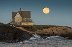 a house sitting on top of a cliff next to the ocean under a full moon