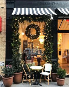 two chairs and a table in front of a store with wreaths on the windows