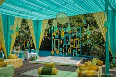 an outdoor wedding setup with yellow and green decorations on the ceiling, blue drapes, and white tablecloths