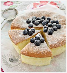 a cake with blueberries and powdered sugar sits on a tablecloth next to a spoon