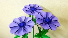 three purple flowers sitting in a green vase on a table next to a white wall