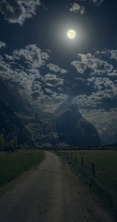 the full moon is shining brightly over a dirt road and grassy field with mountains in the background