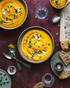 two bowls filled with soup next to some bread and silver spoons on a purple surface