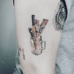 a person with a tattoo on their arm holding a cup filled with paint brushes and pencils