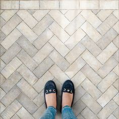 a person wearing blue jeans and black shoes standing on a tile floor with their feet crossed