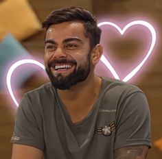 a man with a beard smiles at the camera in front of a heart - shaped neon sign