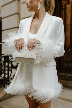 a woman in a white dress and jacket holding a white purse with feathers on it