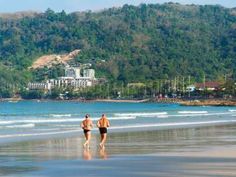 two people are walking on the beach in front of some trees and buildings, with mountains in the background