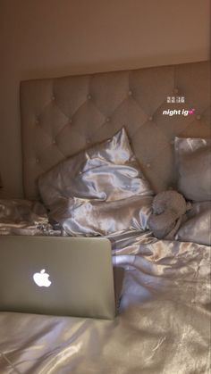 an apple laptop computer sitting on top of a bed with silver sheets and pillow cases