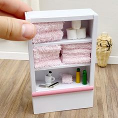 a hand is holding up a miniature shelf with towels and other items in it on a wooden floor