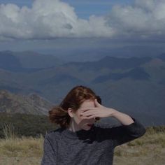 a woman covering her eyes while standing on top of a hill with mountains in the background