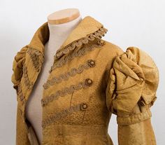 silk damast pelisse 1815 - Made from gold silk damask woven with a pattern of small dots. The bodice, sleeves, and skirt back are lined with brown linen/cotton. Hussar-style Brandenburg trim on the bodice. The collar, cuffs, and bodice front have braided trim. The long narrow sleeves are capped with ruched outher sleeves. The bodice and outer sleeve seams are outlined with corded piping. The skirt is slightly flared in front and full in back. Concealed silk fabric loops and buttons. Regency Costume, Spencer Jacket, Period Fashion