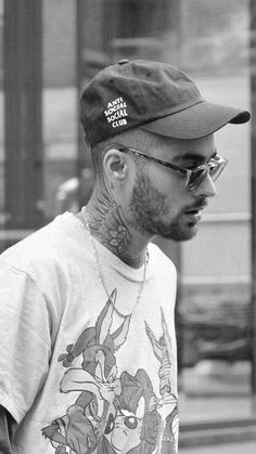 a black and white photo of a man with tattoos on his chest wearing a baseball cap