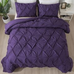 a bed with purple comforter and pillows on top of wooden floor next to potted plant