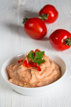 a white bowl filled with hummus next to three red tomatoes on a white surface