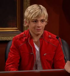 a young man sitting at a table with a microphone in front of him and wearing a red jacket