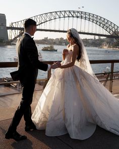 a bride and groom standing on the side of a river with a bridge in the background