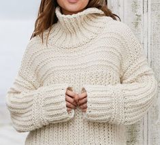 a woman standing in front of a wooden fence wearing a white knitted sweater and hat