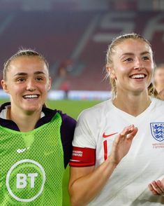 two women standing next to each other in front of a soccer field with people watching