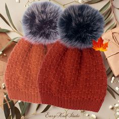 two knitted hats with faux fur pom - poms sit on top of each other