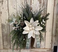 a white flower arrangement with pine cones and greenery in front of a wooden wall