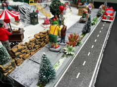 a toy train set with people and trees on the tracks, in front of an open road