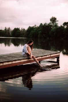 a woman sitting on the end of a dock over water with trees in the background