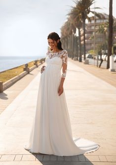 a pregnant woman in a white dress standing on the sidewalk by the ocean with palm trees