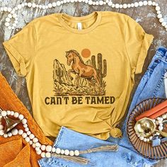 "Can't Be Tamed " Running Horse Design Available in Mustard, Mauve, & Cream Super Soft & Stretchy Cotton Tee Unisex Cut for Men or Women ~ Runs Roomy ~ If You Want A More Fitted Tee We Recommend You Size Down Women's Sizing Recommendations Small 0/2/4 Medium 4/6/8 Large 8/10/12 XLarge 12/14/16 2XLarge 16/18/20 3XLarge 20/22/24 ***These Are PRINTED/MADE TO ORDER! Please Allow 7-10 Business Days To Ship. If You Order Multiple Items, These Will Be Shipping Out SEPARATELY From Your Order. Western Shopping, Western Tee Shirts, Western Inspiration, Can't Be Tamed, Staging Ideas, Western Clothes, Western Tee, Western Graphic Tees, Country Clothing