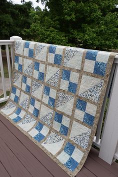 a blue and white quilt is hanging on a porch railing with trees in the background